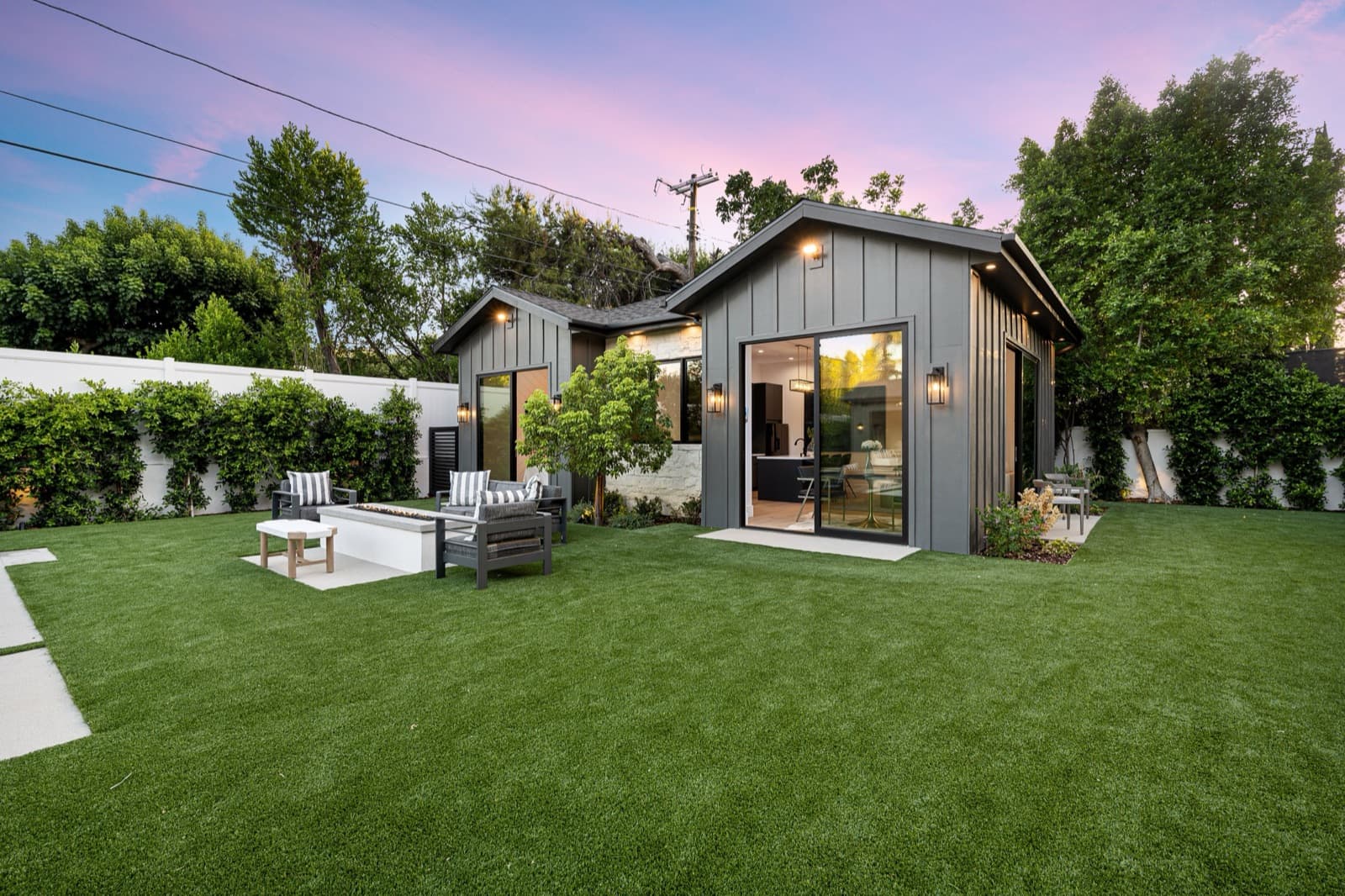 A detached accessory dwelling unit at dusk, with a gable roof, board-and-batten siding, and sliding glass doors opening onto a patio and lawn.